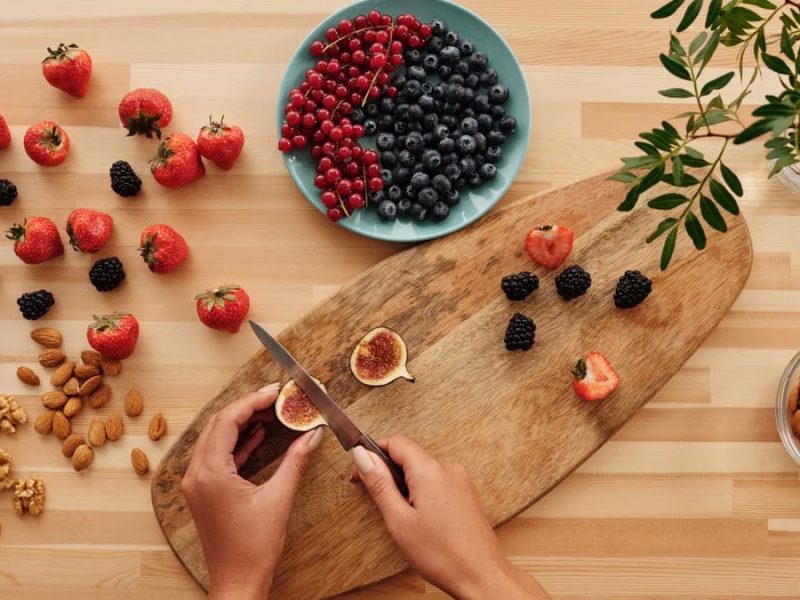 Overhead shot of fresh fruits, nuts, and hands preparing ingredients on a wooden board.